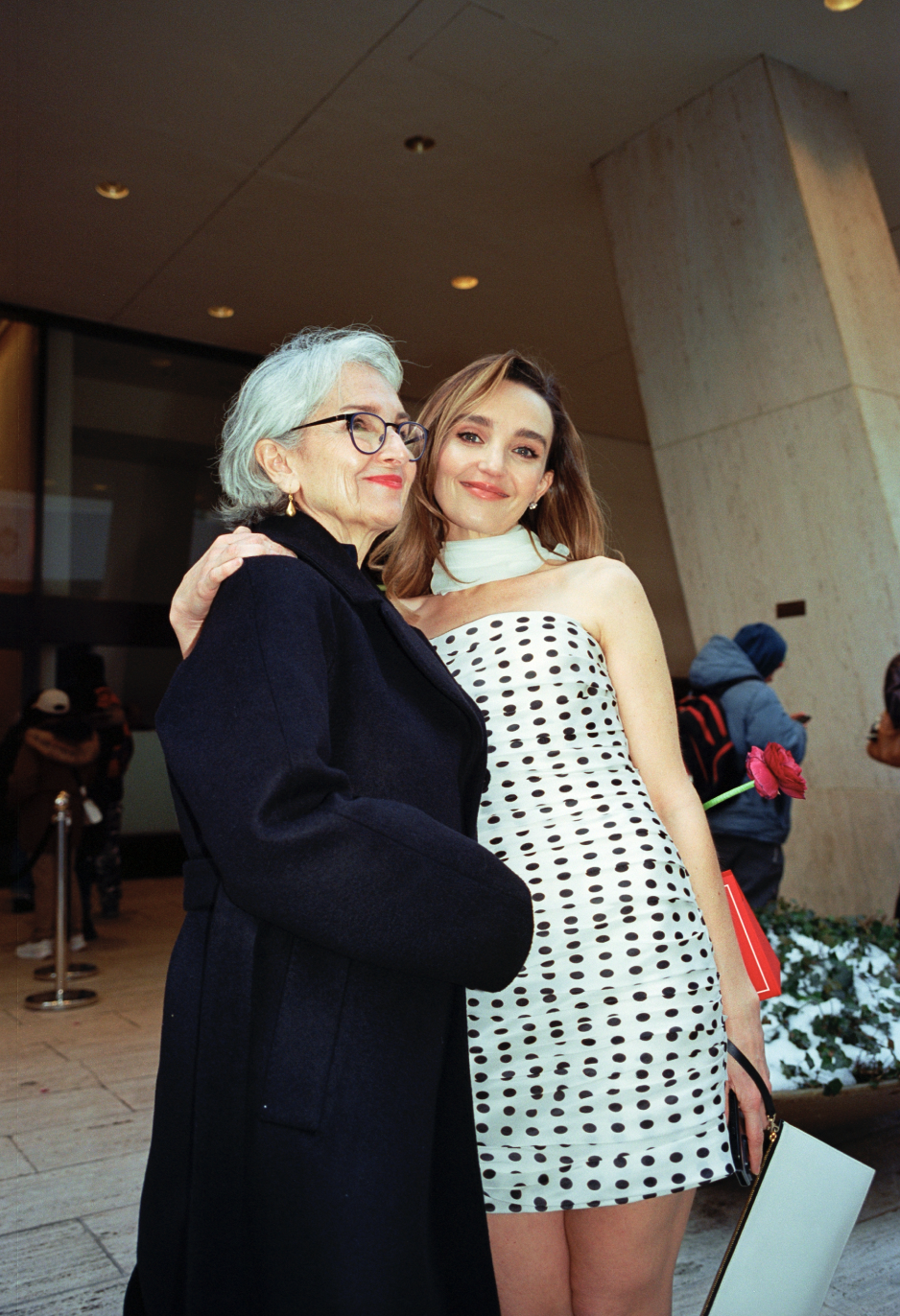 Two women standing close together indoors; one older with gray hair, glasses, and wearing a dark coat, and one younger with light brown hair in a strapless polka dot dress, holding a red rose.