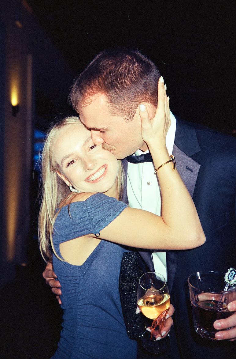 A couple in formal attire sharing an affectionate kiss and hug at a celebration, with the woman holding a glass of champagne.