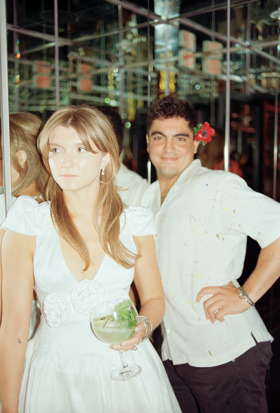 A woman in a white dress holding a cocktail, and a man with a pink flower in his hair, smiling at a social gathering, with mirrors and reflections in the background.