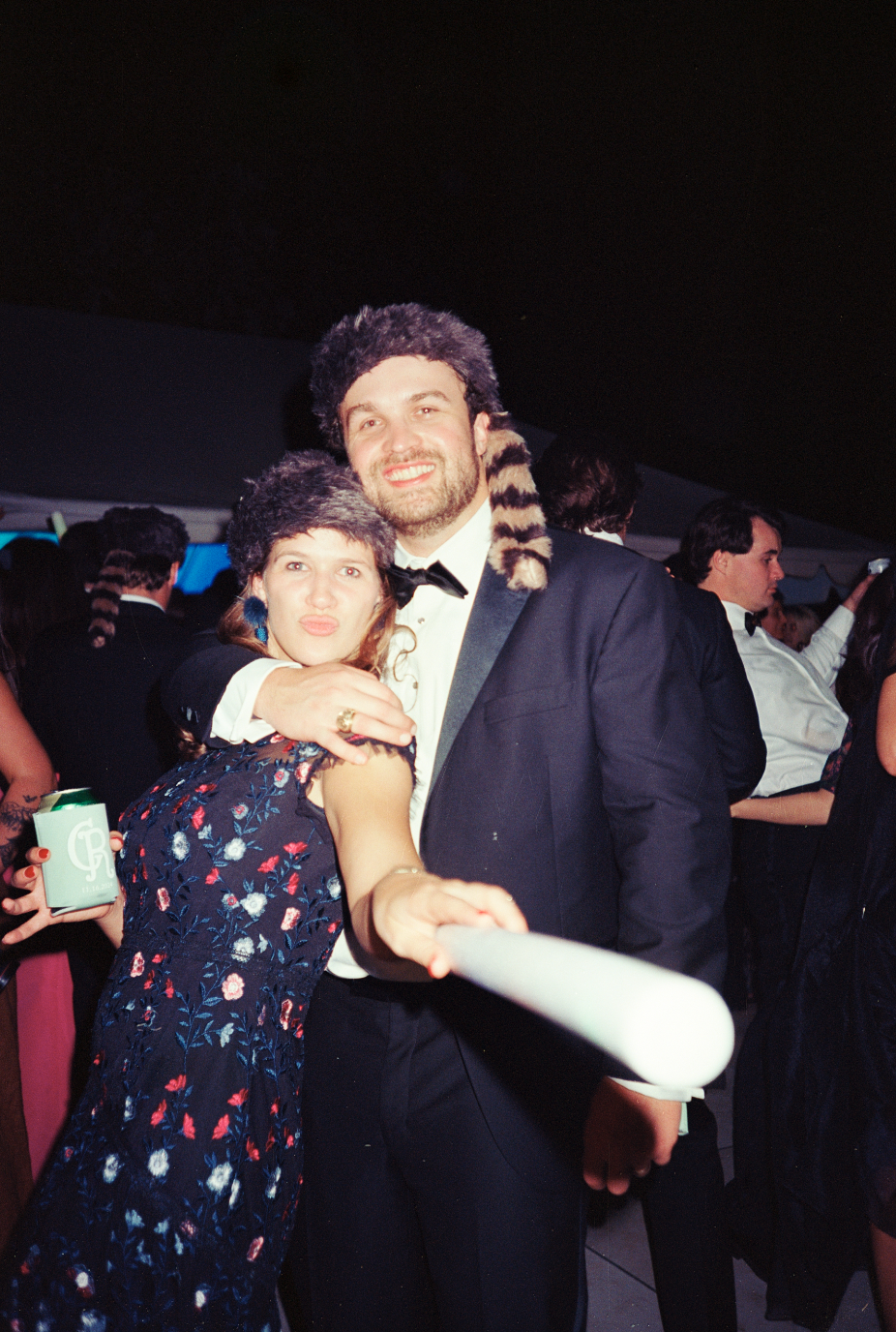A woman and man dressed in formal attire at a party, taking a selfie together. The woman is wearing a floral dress and a fuzzy hat, and the man is in a tuxedo with a bow tie, holding a selfie stick. The woman is making a playful pout, and the man has