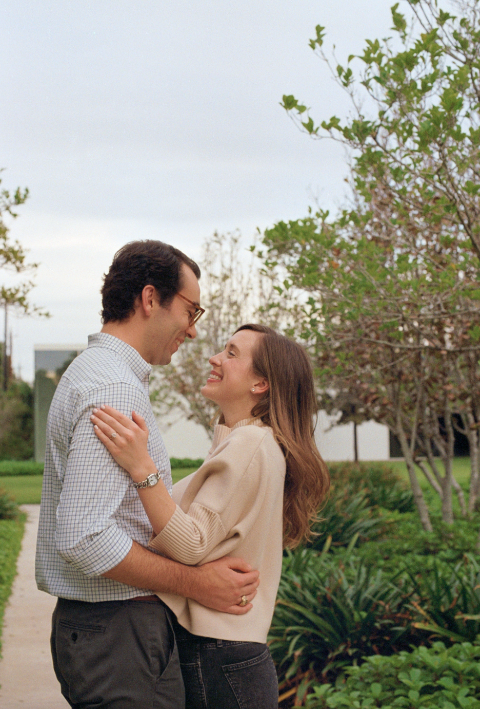 A couple happily embracing outdoors on a cloudy day, surrounded by green shrubs and trees.