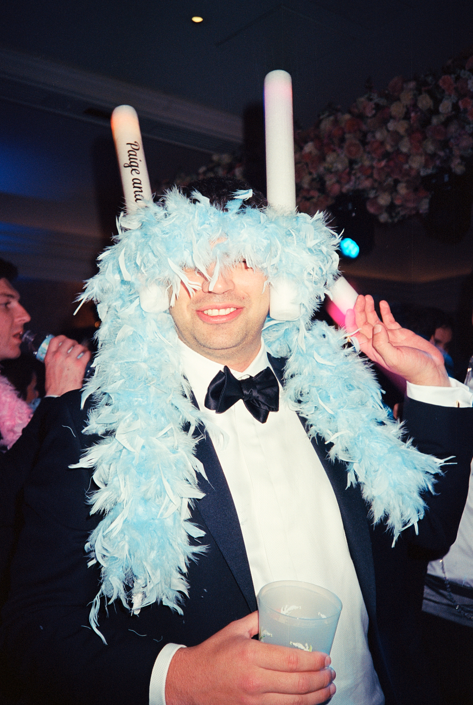 Man dressed in a tuxedo with a feathered blue hat, holding a drink, celebrating at a party.