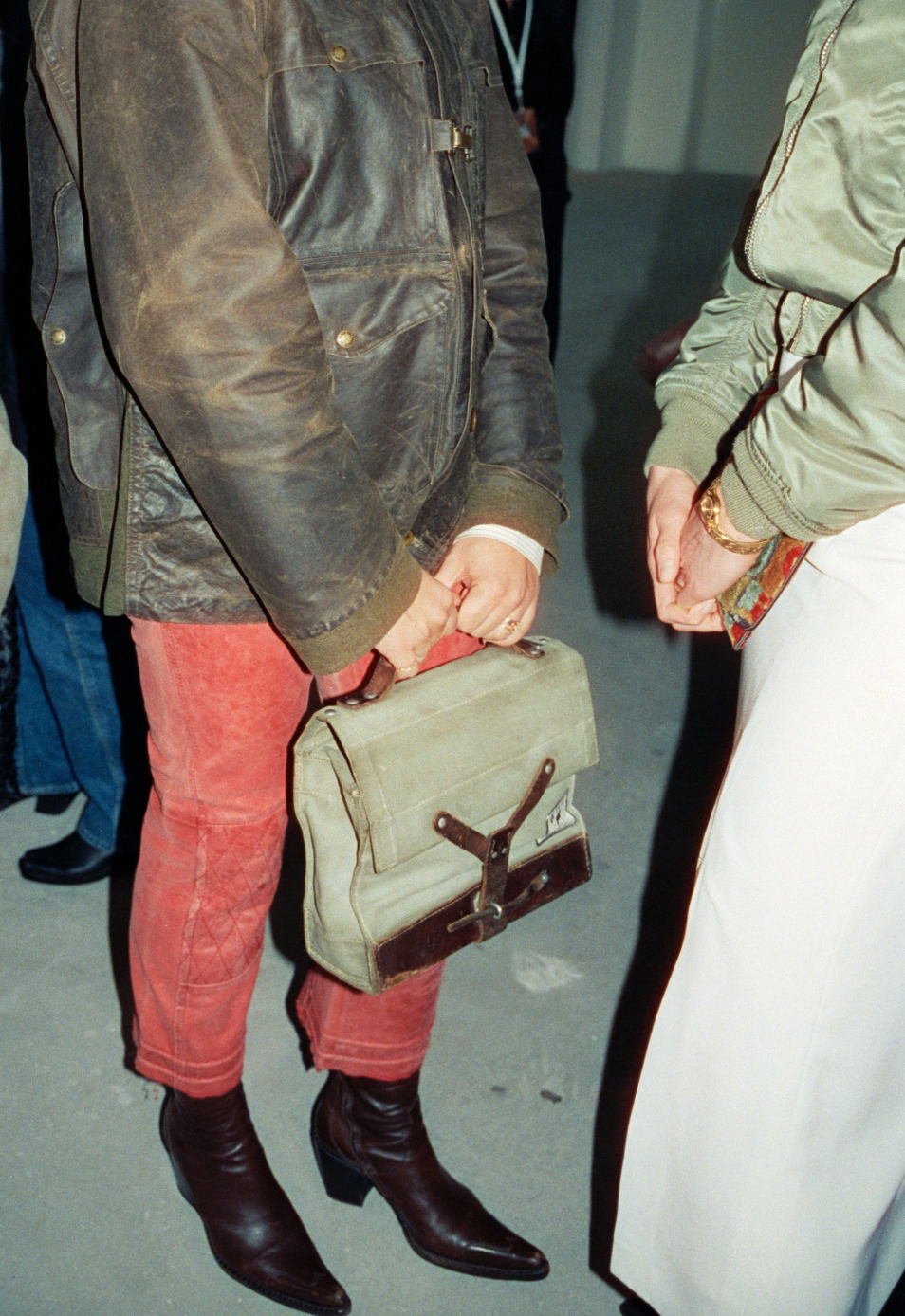Two people standing and talking, dressed in vintage clothing, with one holding a beige bag and wearing dark brown boots.