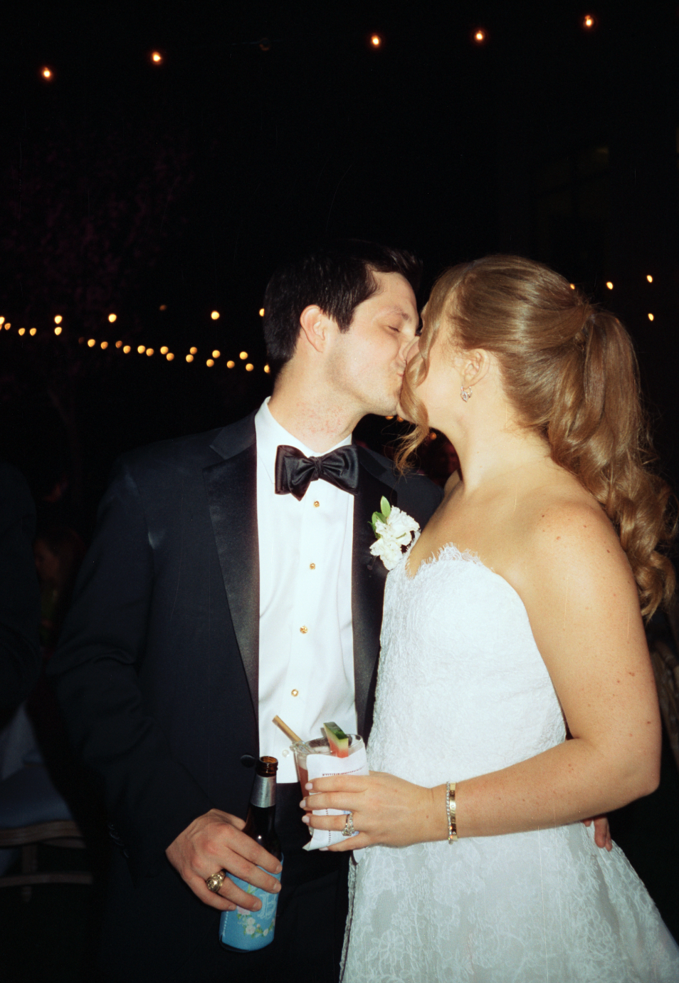 A newlywed couple sharing a kiss at night, dressed in wedding attire, with string lights in the background.