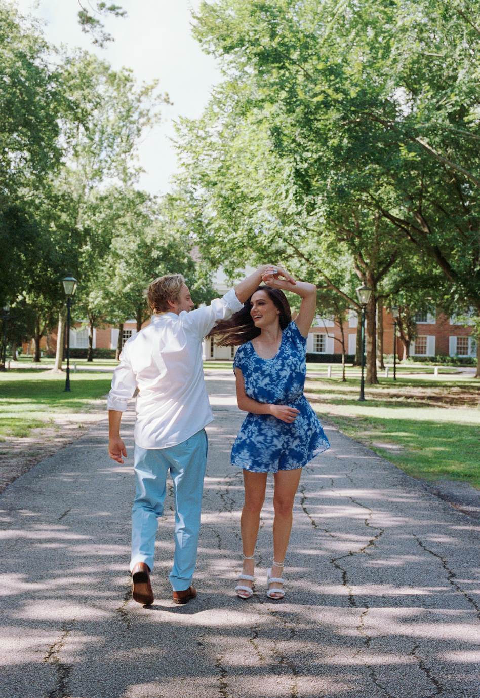A young couple dancing on a cracked asphalt path in a park, with green trees and school buildings in the background, under a sunny sky.