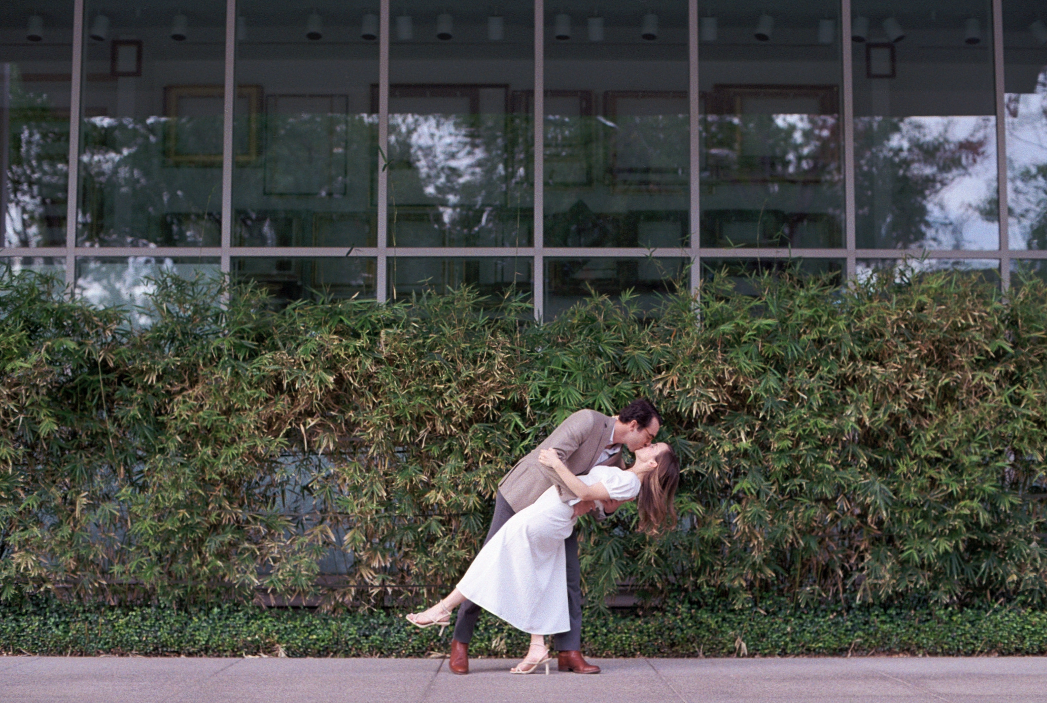 A man and woman engaged in a romantic kiss outdoors, with the man dipping the woman backward in front of a large brick wall with a window. The woman is wearing a white dress and heels, and the man is in a beige blazer, dark pants, and brown boots.