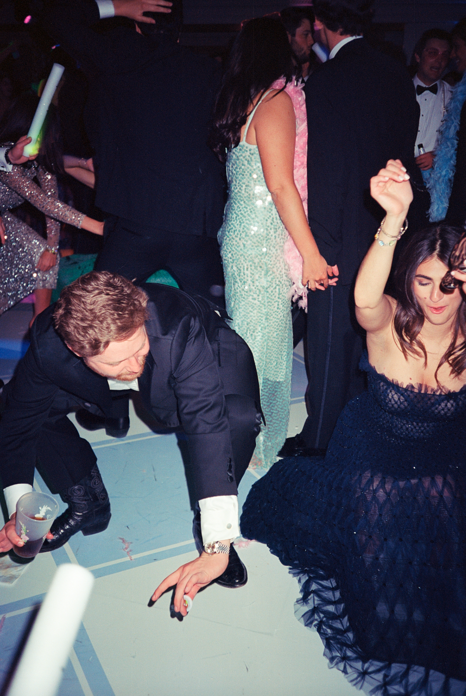 People dancing and socializing at a party or celebration, dressed in formal and glamorous attire, with one person crouching on the floor holding a drink.