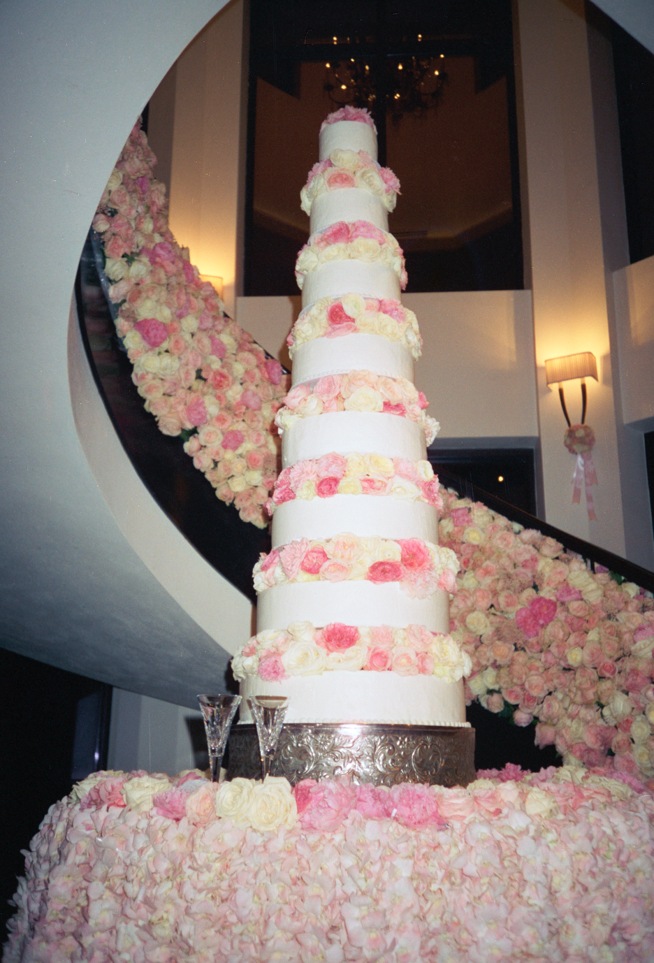 A tall, tiered wedding cake decorated with pink, white, and peach flowers, placed on a table surrounded by pink and white flower petals. The background features a staircase adorned with flowers, and the setting appears to be inside a venue with warm 