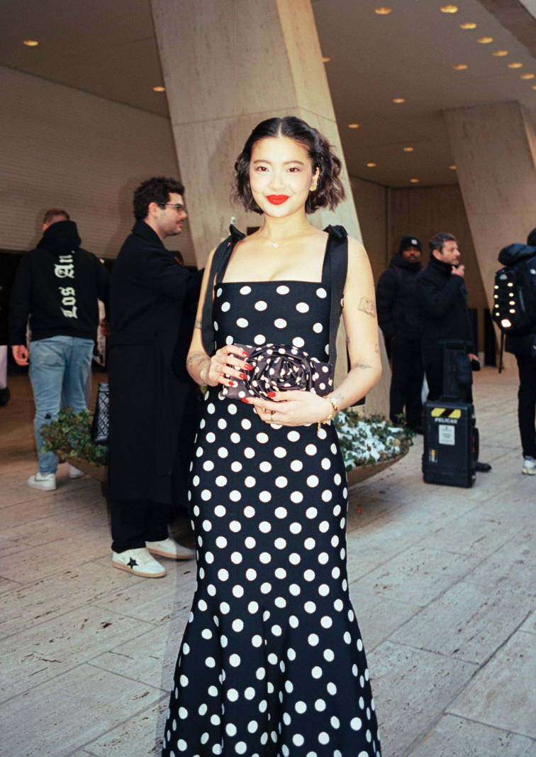 A woman with short wavy hair wearing a black strapless polka dot dress with red lipstick standing outdoors with people in the background.