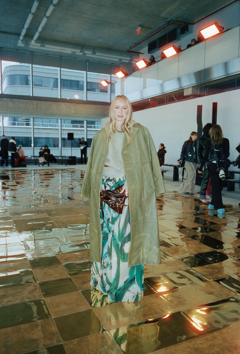A woman with long blonde hair in a light green coat and colorful patterned pants stands inside a modern airport terminal with reflective tile floors and large windows.