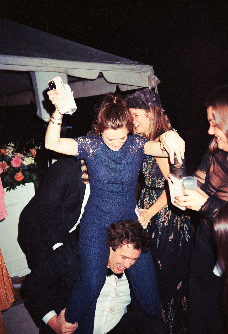 Group of people celebrating and having fun at a party with a woman sitting on a man's shoulders, using her phone to take pictures, and others smiling and holding drinks.