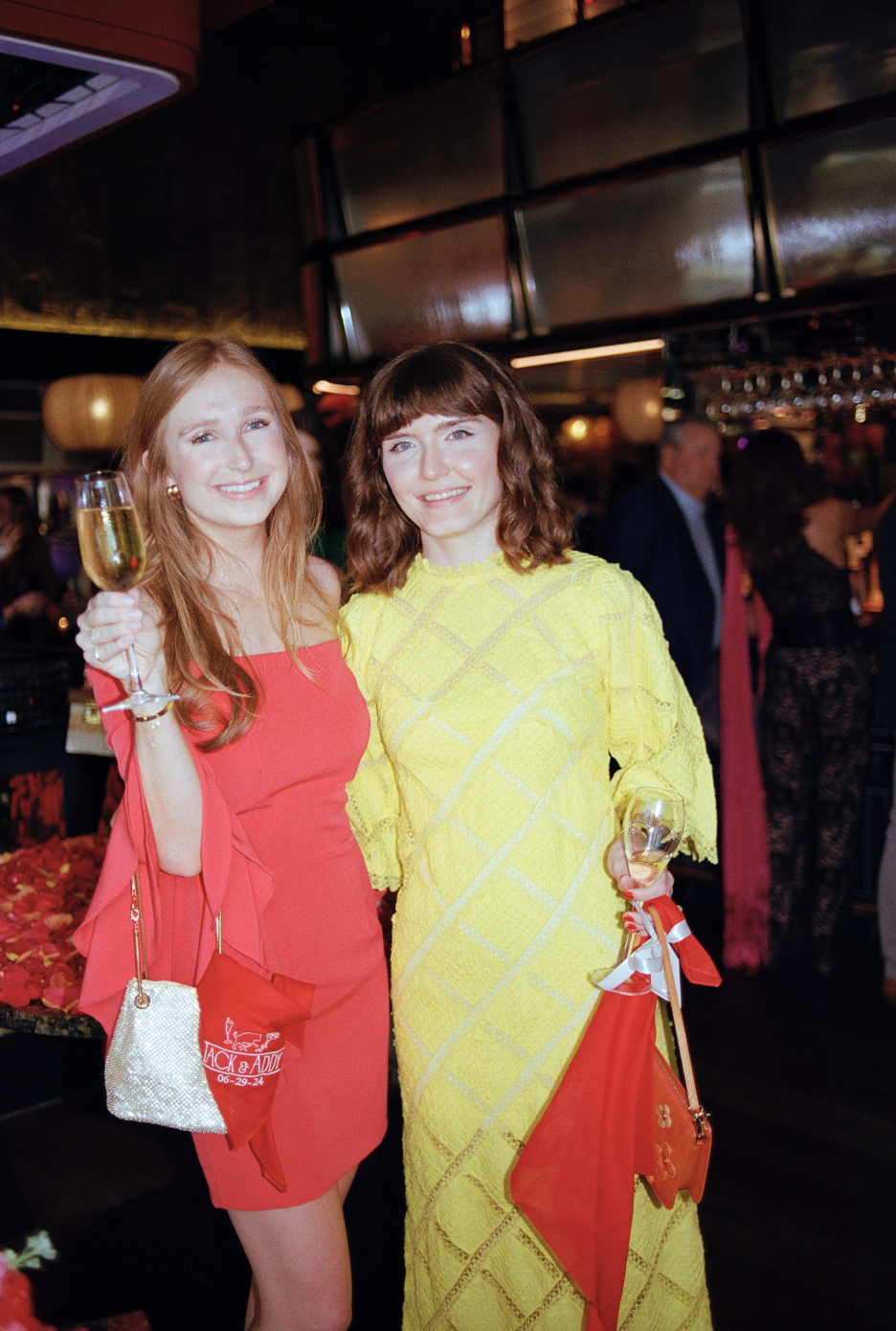 Two women smiling at a celebratory event, holding glasses of champagne, with one in a red dress and the other in a yellow dress, in a dimly lit social setting.