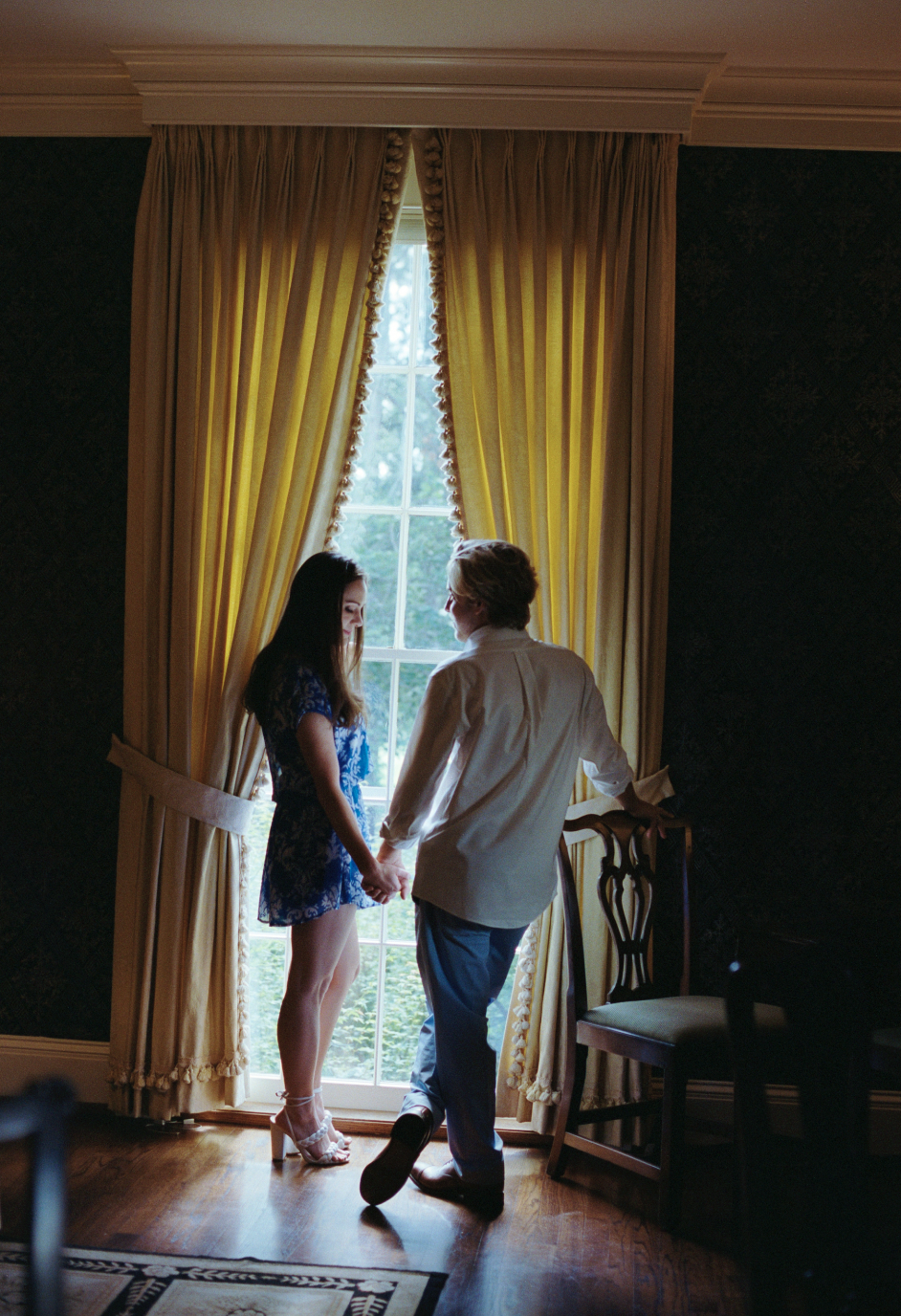 A woman in a blue lace dress and high heels holding hands with a man in a white shirt and dark pants, standing by a window with yellow curtains, indoors.