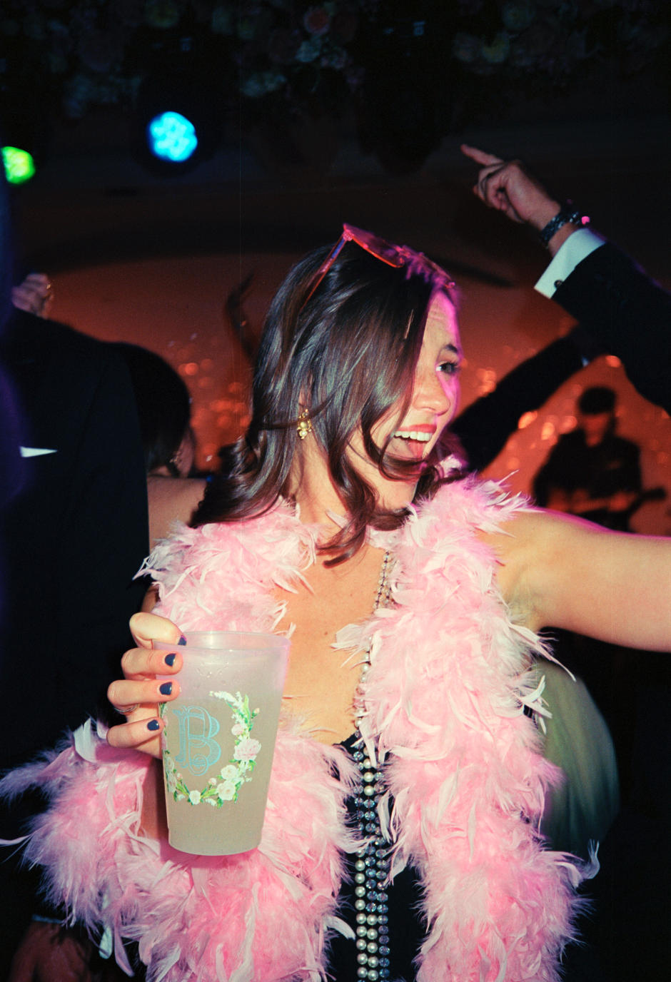 Woman dancing at a party wearing a pink feather boa, holding a drink, and smiling.