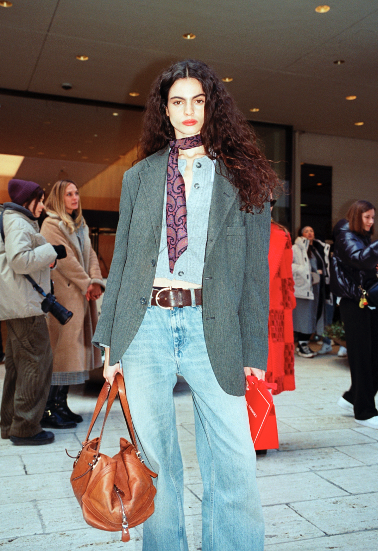 Fashionably dressed woman with curly hair in a blazer, jeans, and scarf, standing in front of a group of people in an indoor public space.