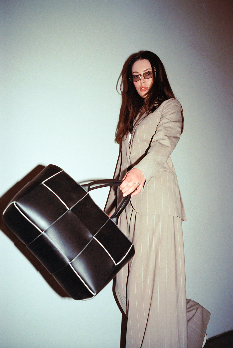 Woman with long dark hair wearing sunglasses and a beige pinstripe suit holding a large black and white grid-patterned bag.