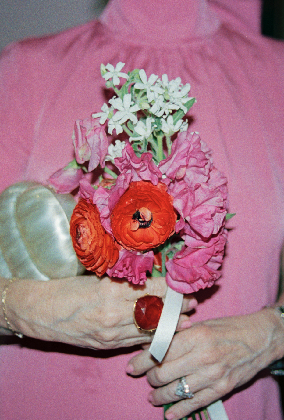 A person in a pink dress holding a bouquet of pink, white, and red flowers, with a large red ring on their finger and a pearl bracelet on their wrist.