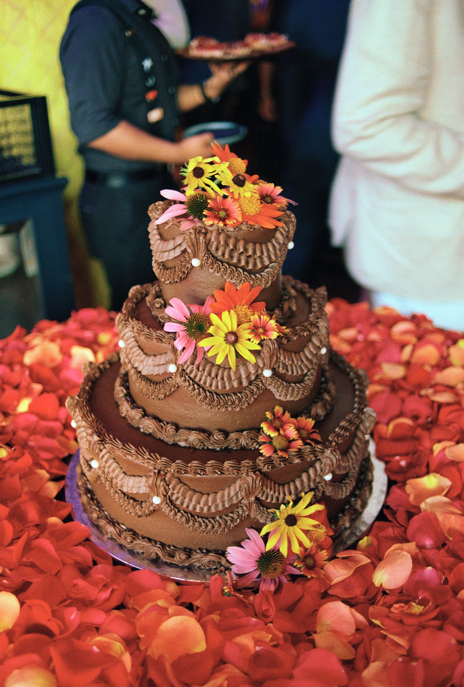 A multi-tiered chocolate cake decorated with colorful flowers, surrounded by rose petals, at a celebration event.
