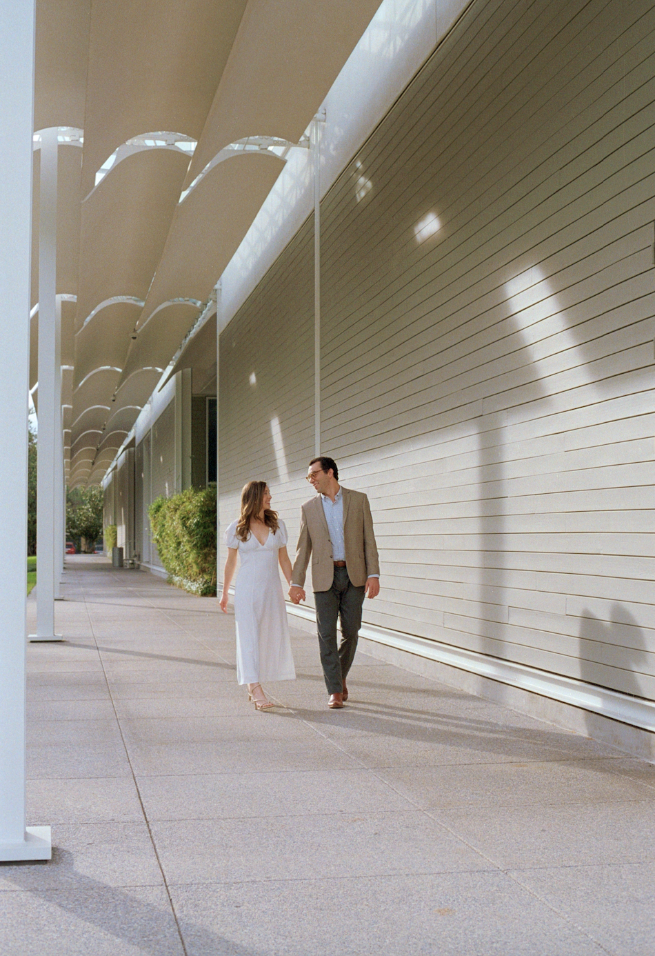 A couple walking hand in hand outside a modern building with architectural sun shades on the roof.