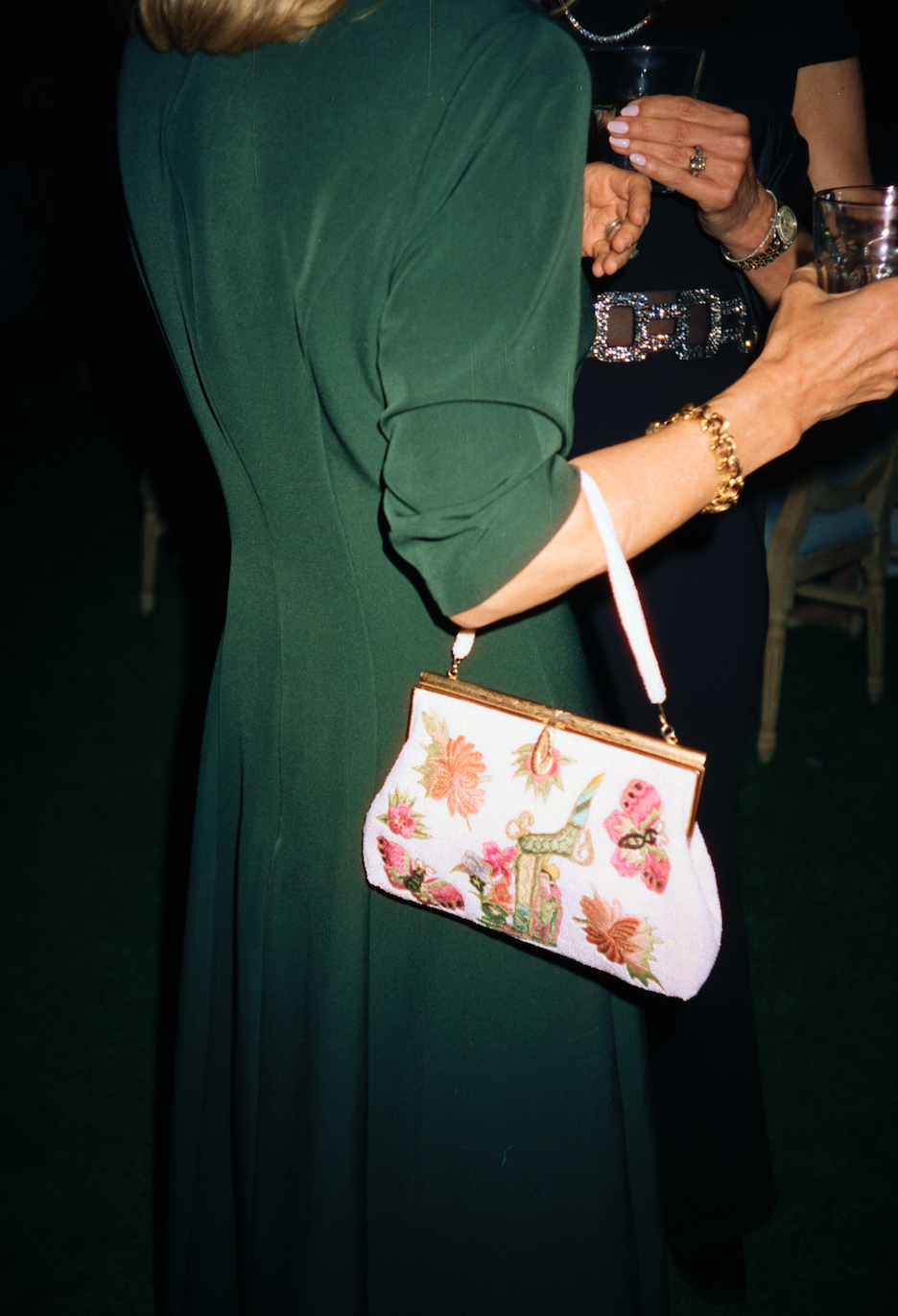A woman in a green dress holding a floral embroidered clutch purse and a glass of drink at a social event.