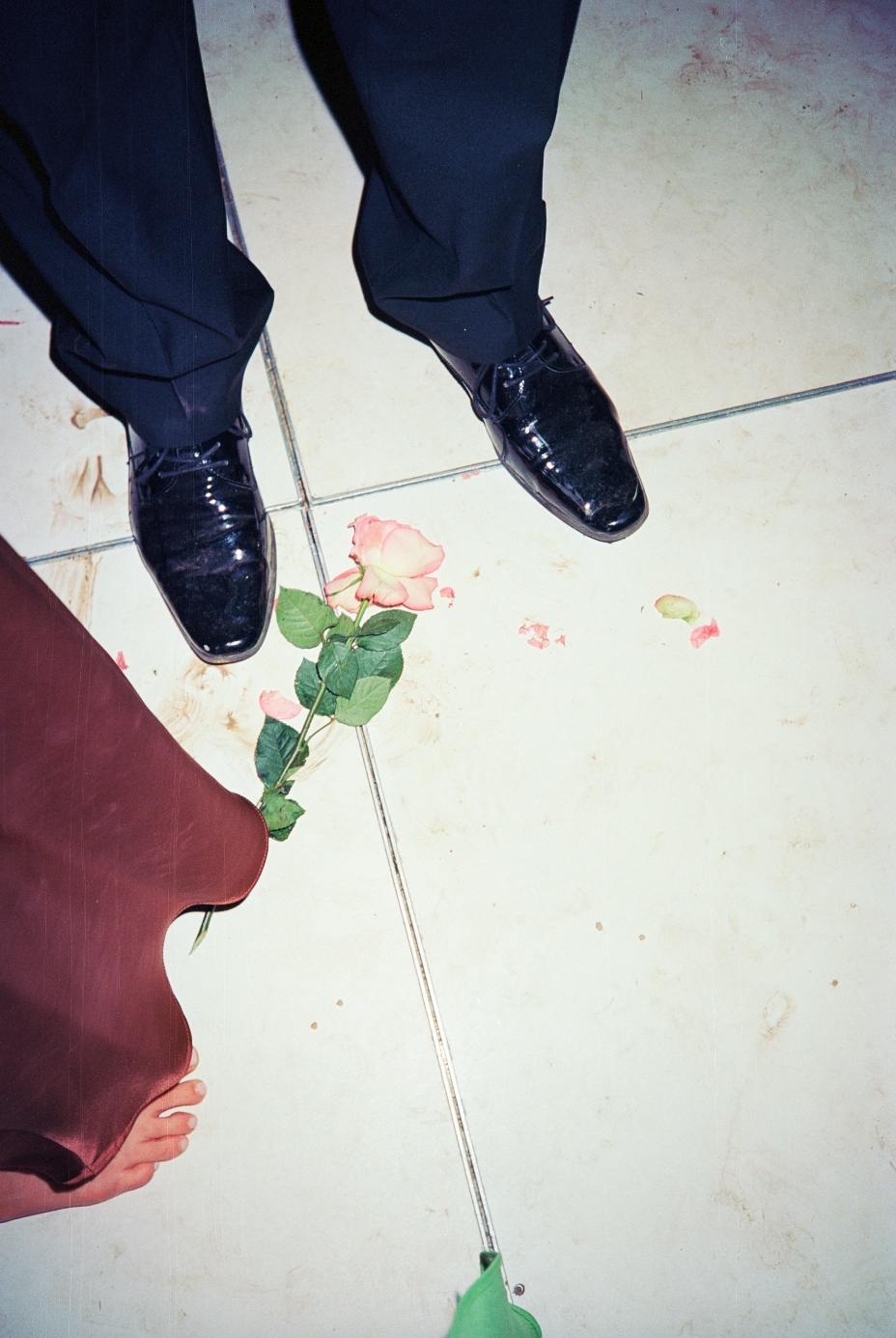 Top-down view of a pair of black shiny dress shoes and black pants, with a pink rose on the tiled floor and a person's bare foot near the edge of a maroon fabric or clothing item, and a green object connected to a string or cable.