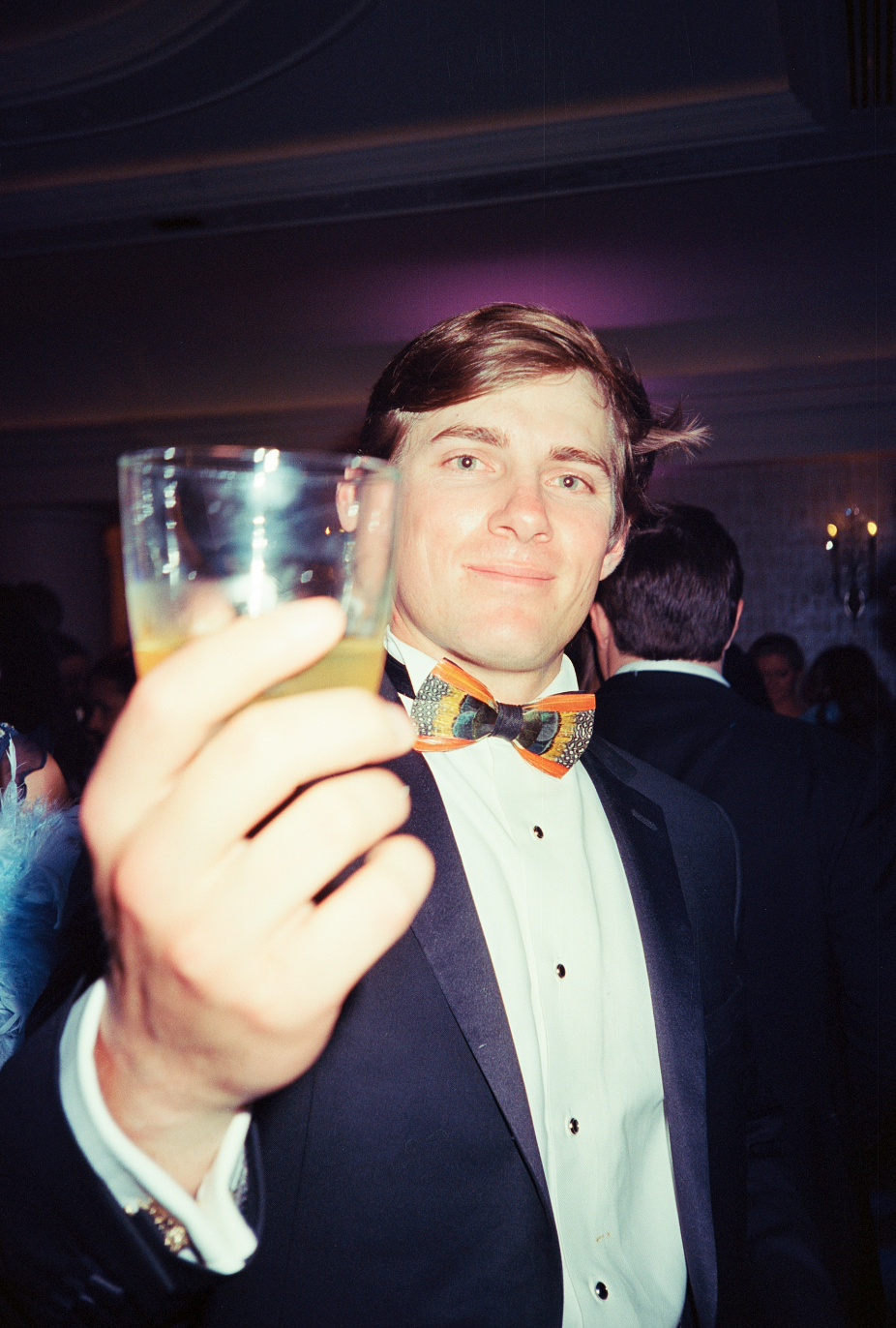A young man in a tuxedo and colorful bowtie holding a glass of drink at a party or celebration, with other guests in the background.