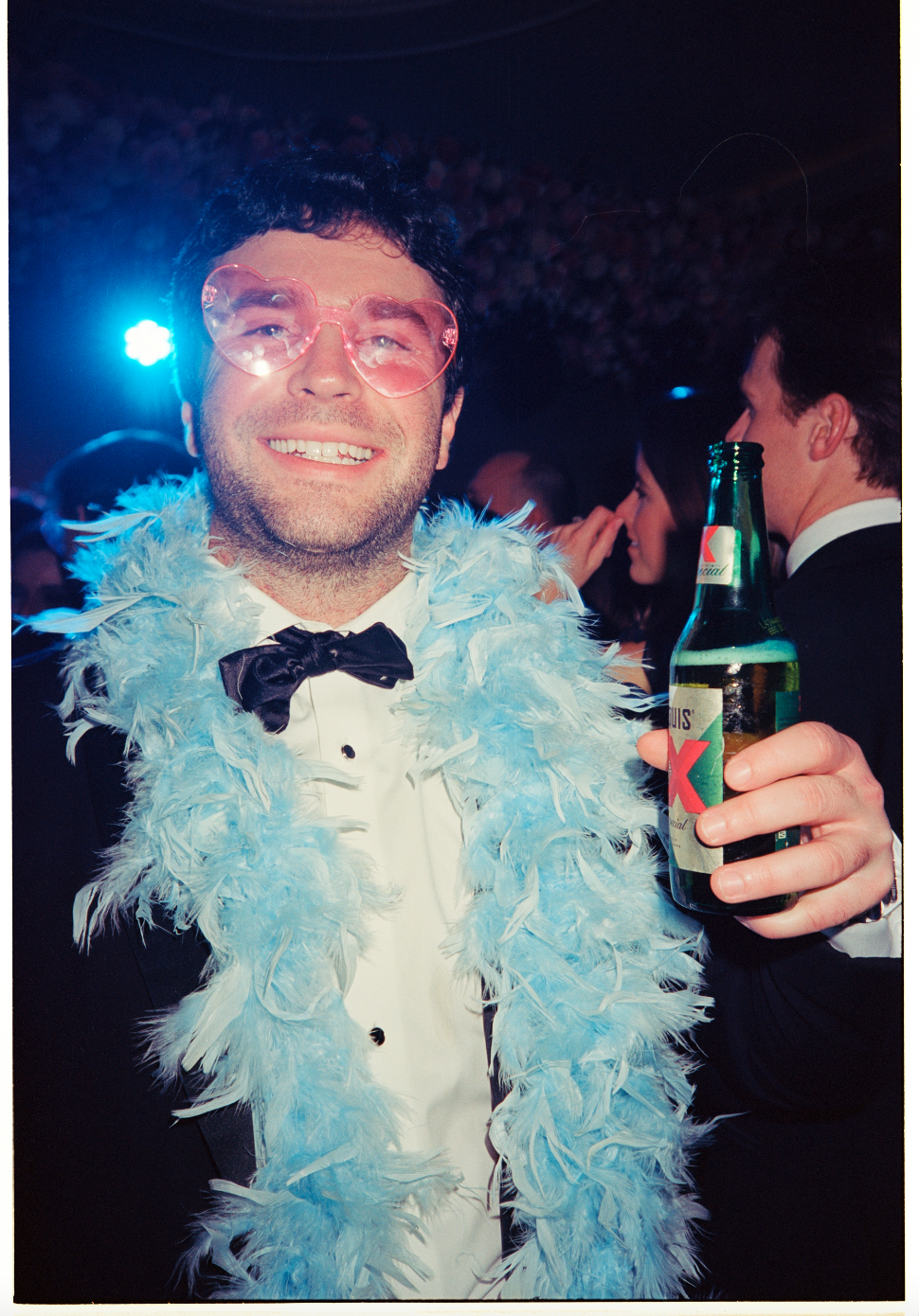 A man at a party wearing pink heart-shaped glasses, a feather boa, a tuxedo, and holding a bottle of beer, smiling at the camera.