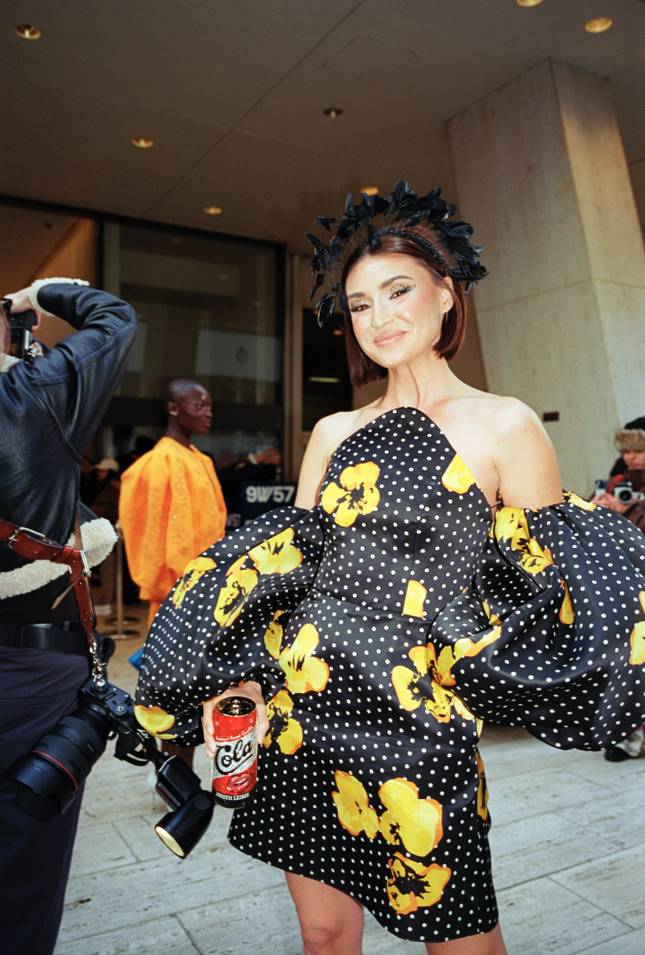 A woman in a black and yellow floral and polka dot dress with puffed sleeves, wearing a black feathered headpiece, holding a red soda can, smiling at the camera at an indoor event.