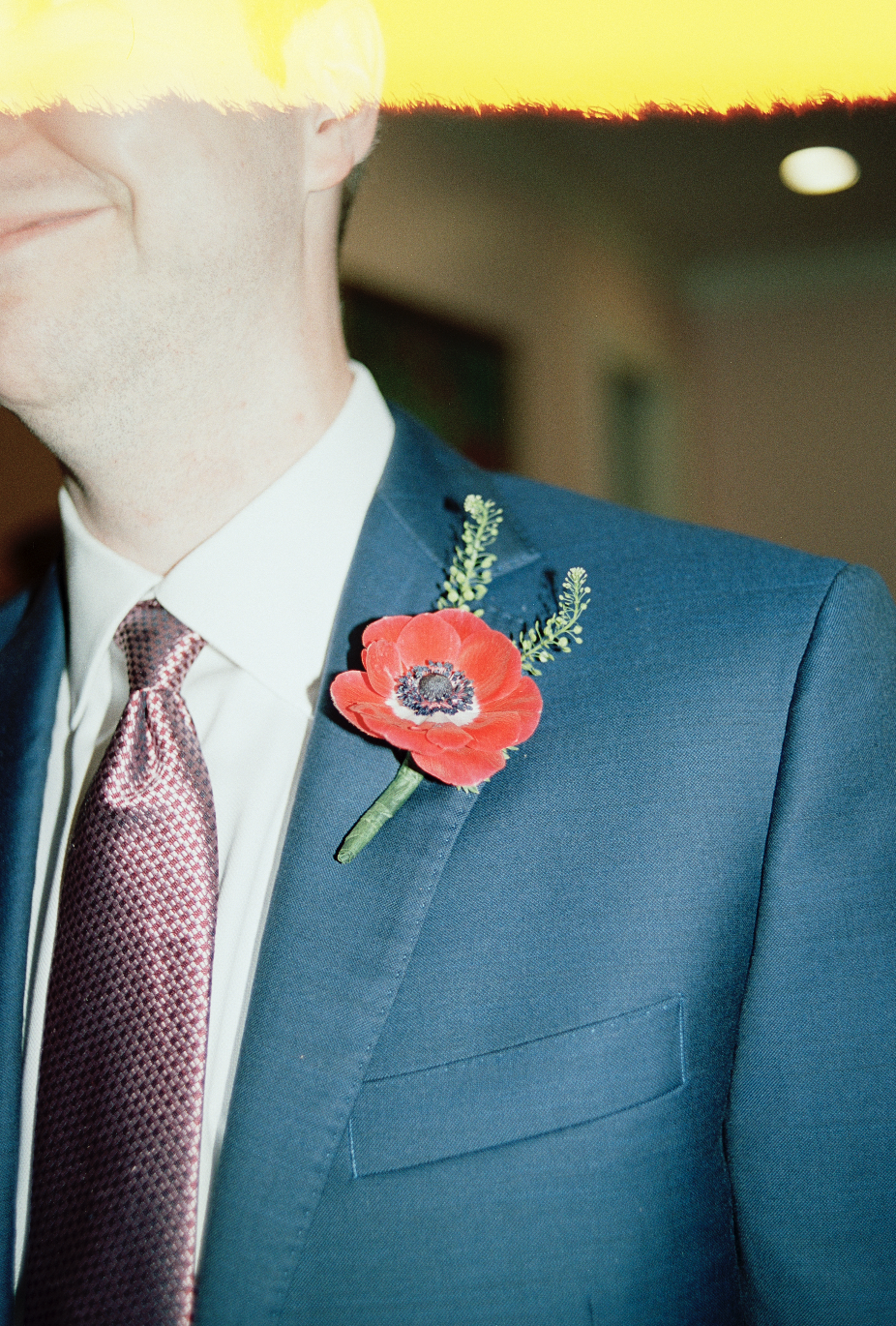 Close-up of a man in a blue suit with a red flower boutonniere on his lapel, wearing a tie and white shirt, inside a building.