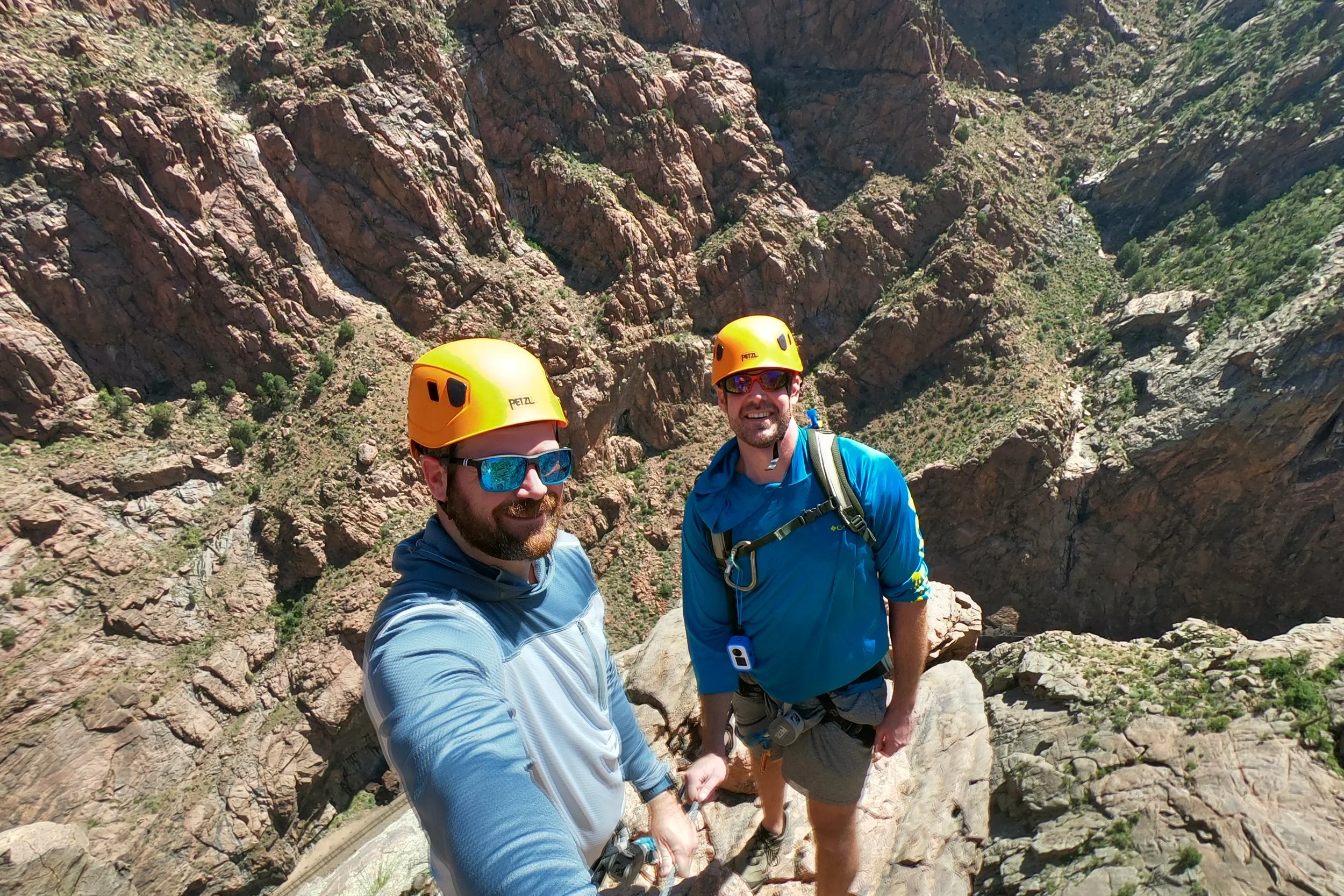 Two men in blue outdoor gear and yellow helmets hiking in rocky canyon.