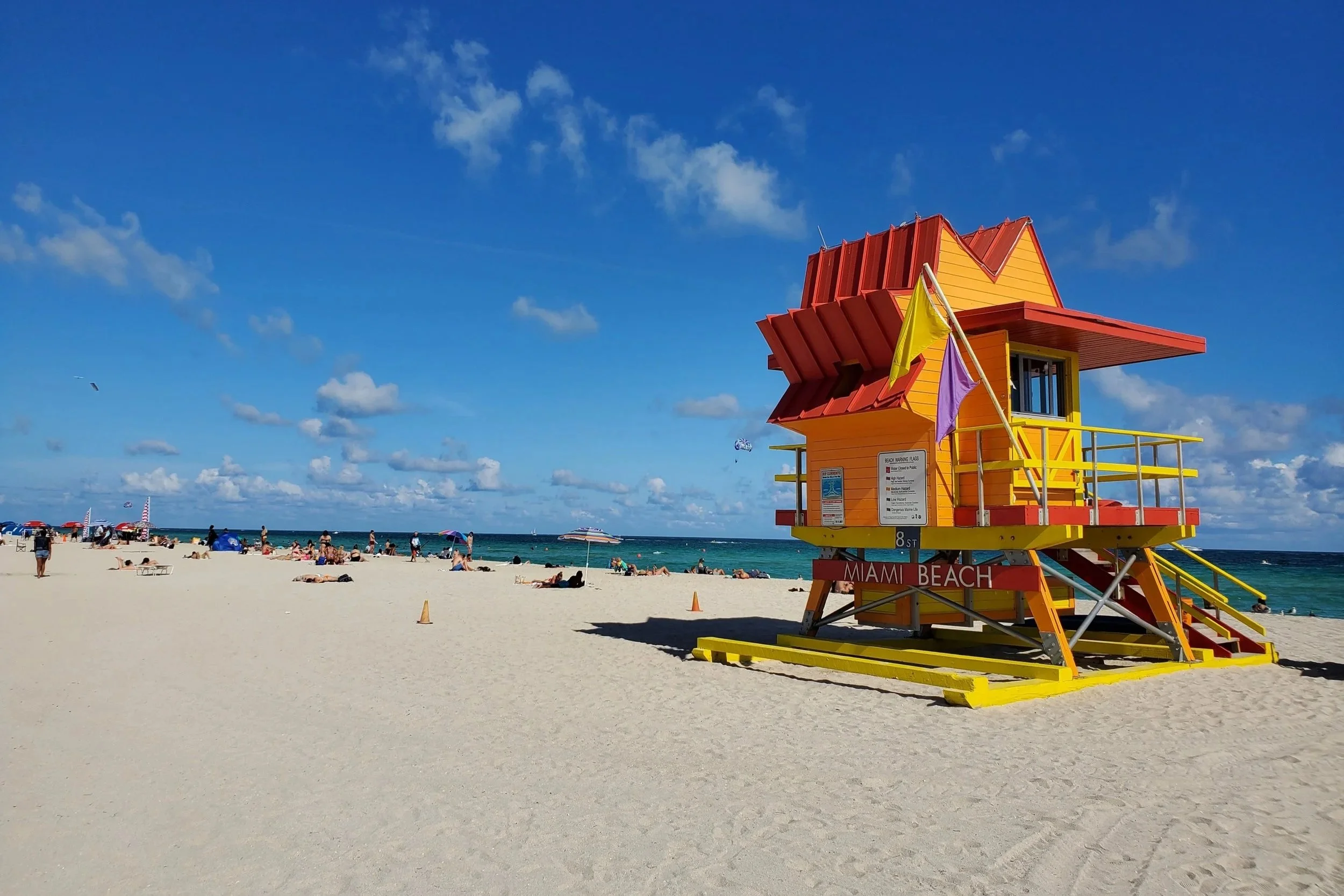 Colorful lifeguard tower on Miami Beach with people relaxing on the sandy beach and blue sky with clouds in the background.
