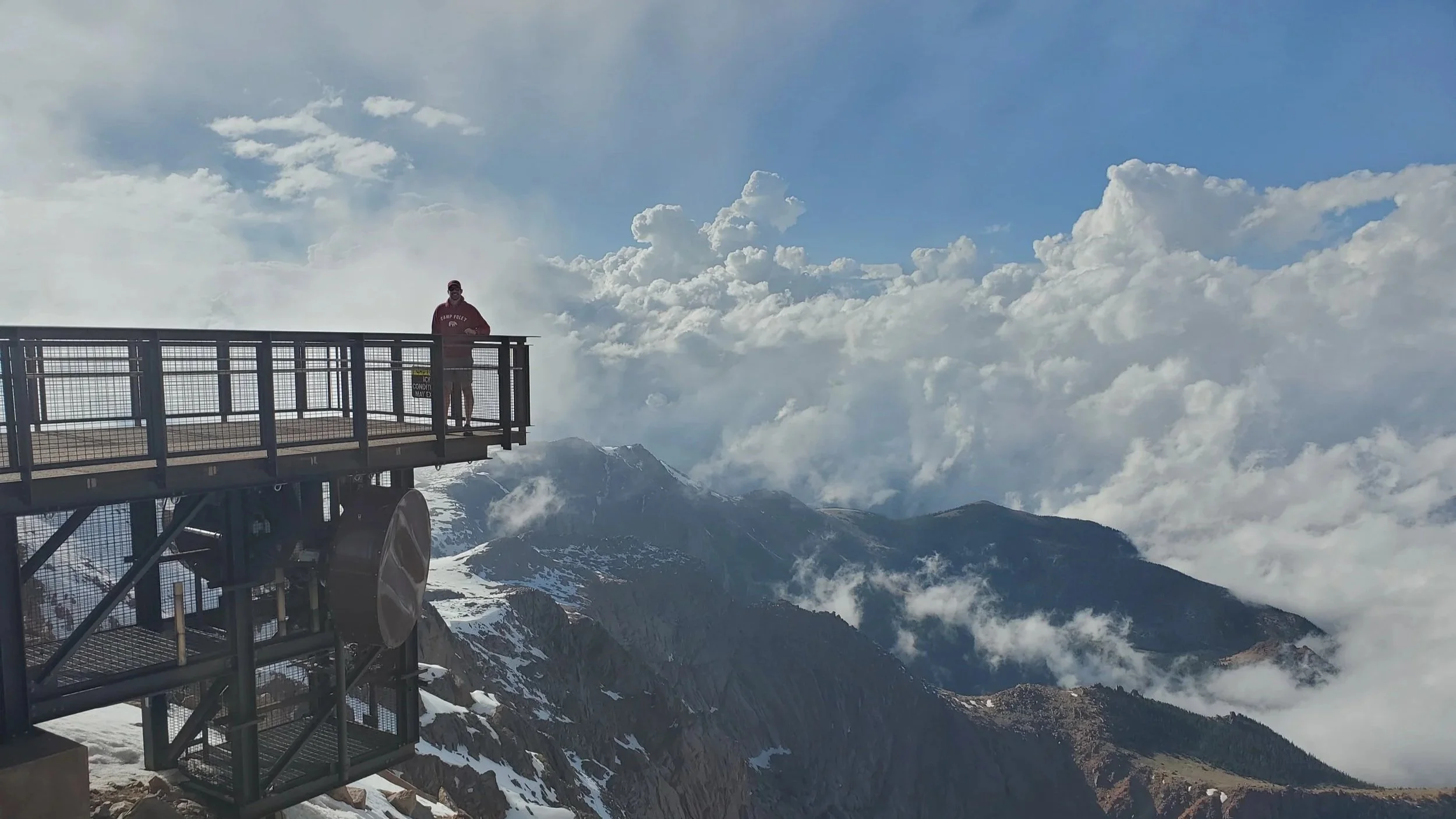 A person standing on a metal observation deck at a mountain summit, with snow-capped peaks and clouds in the background. Pike's Peak, Colorado Springs