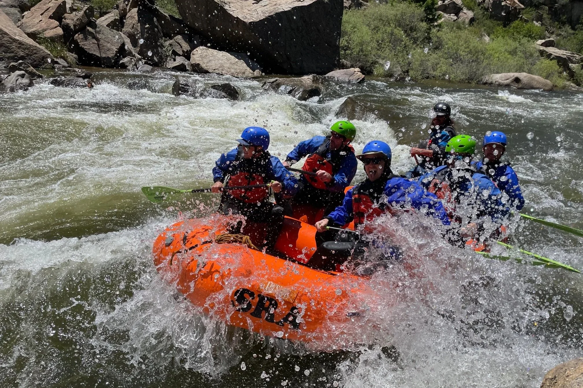 A group of people white water rafting in a river, wearing helmets and life jackets, with water splashing around them.