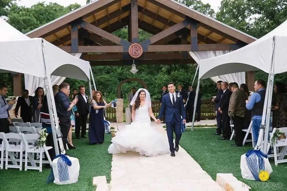 Bride and groom walking hand in hand down the wedding aisle under a pavilion, surrounded by guests.