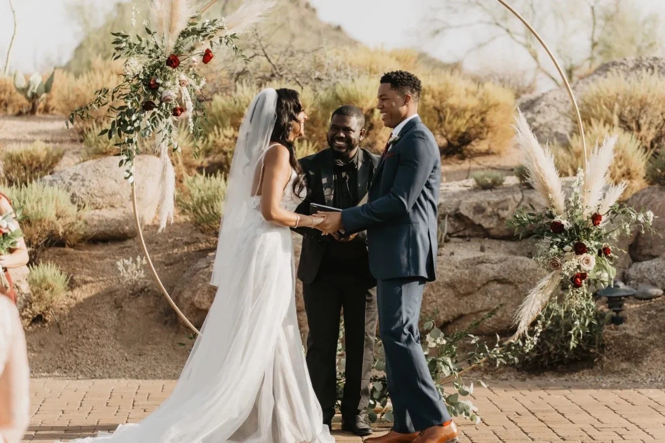 A couple getting married outdoors during daytime, holding hands and looking at each other, with an officiant standing between them. The scene features a decorative floral arch and desert-like landscape with rocks and bushes in the background.