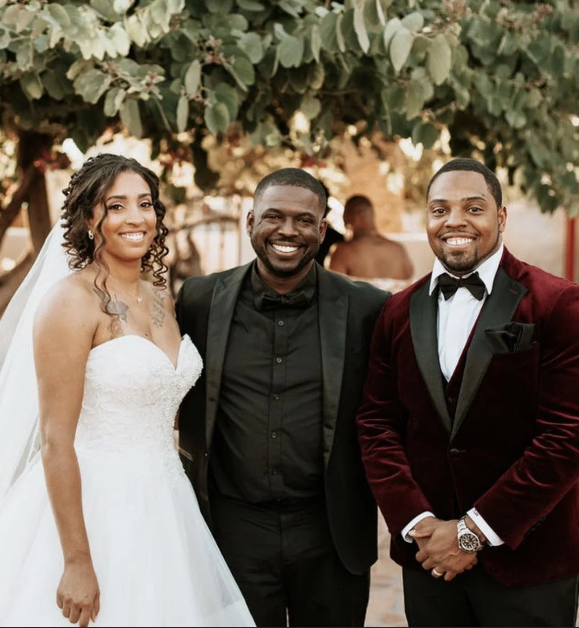 A bride and two men smiling at a wedding celebration outdoors, with a lush green tree in the background.