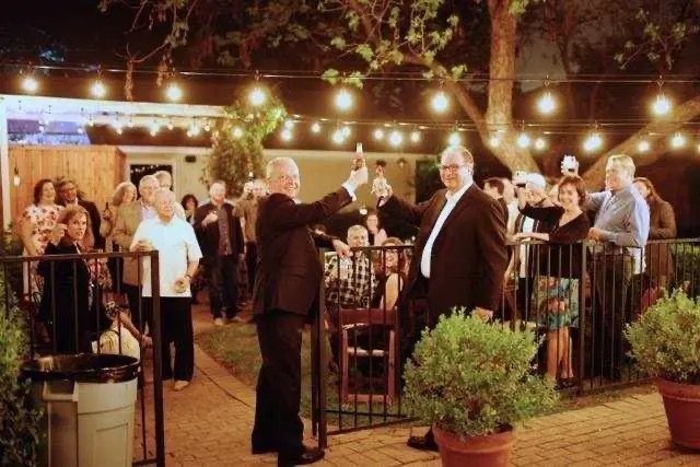 Two men in suits toasting at an outdoor evening gathering, with people watching and string lights overhead.