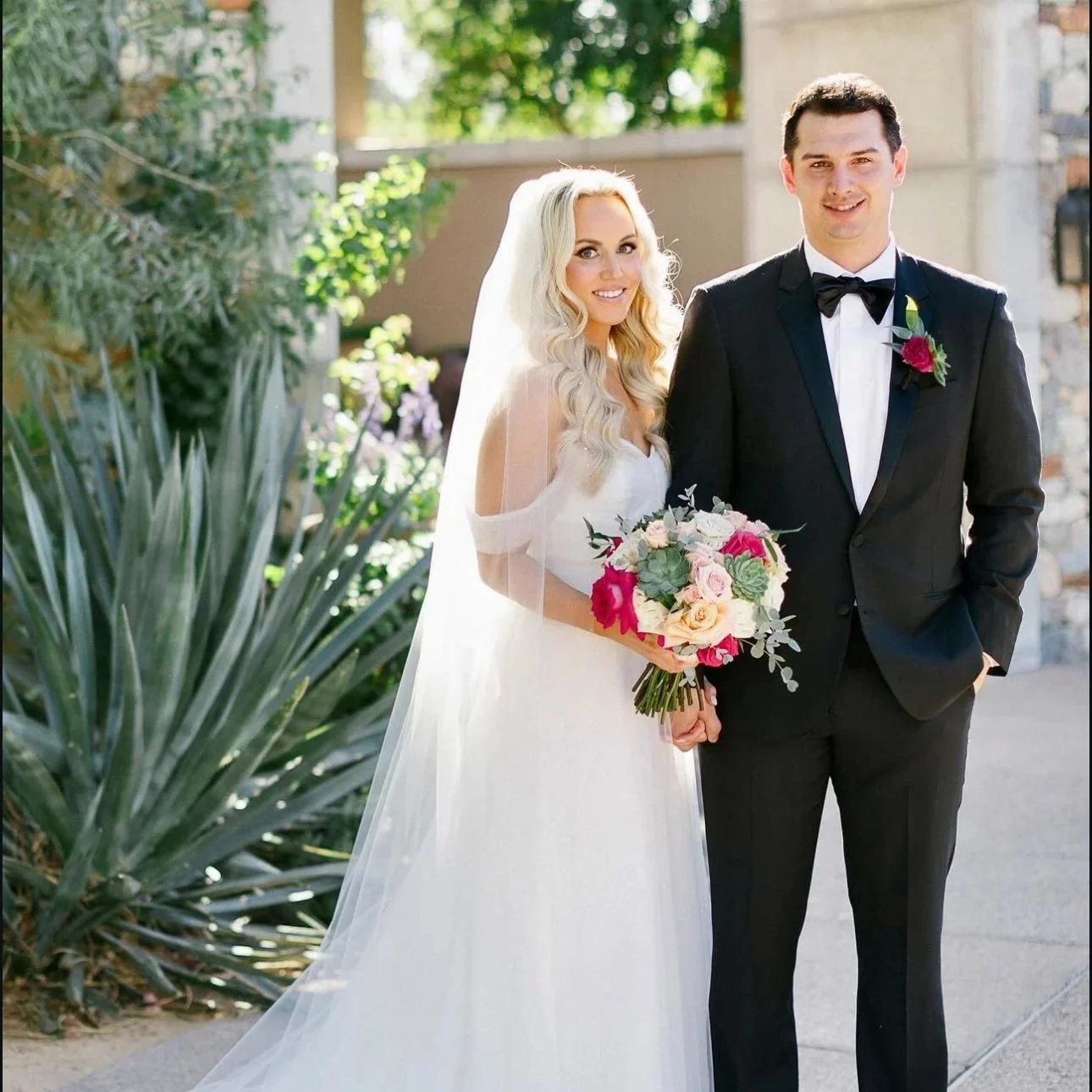 A bride and groom standing outdoors on their wedding day, smiling, with the bride holding a bouquet of pink, white, and lavender flowers, and the groom dressed in a black tuxedo with a red boutonniere.