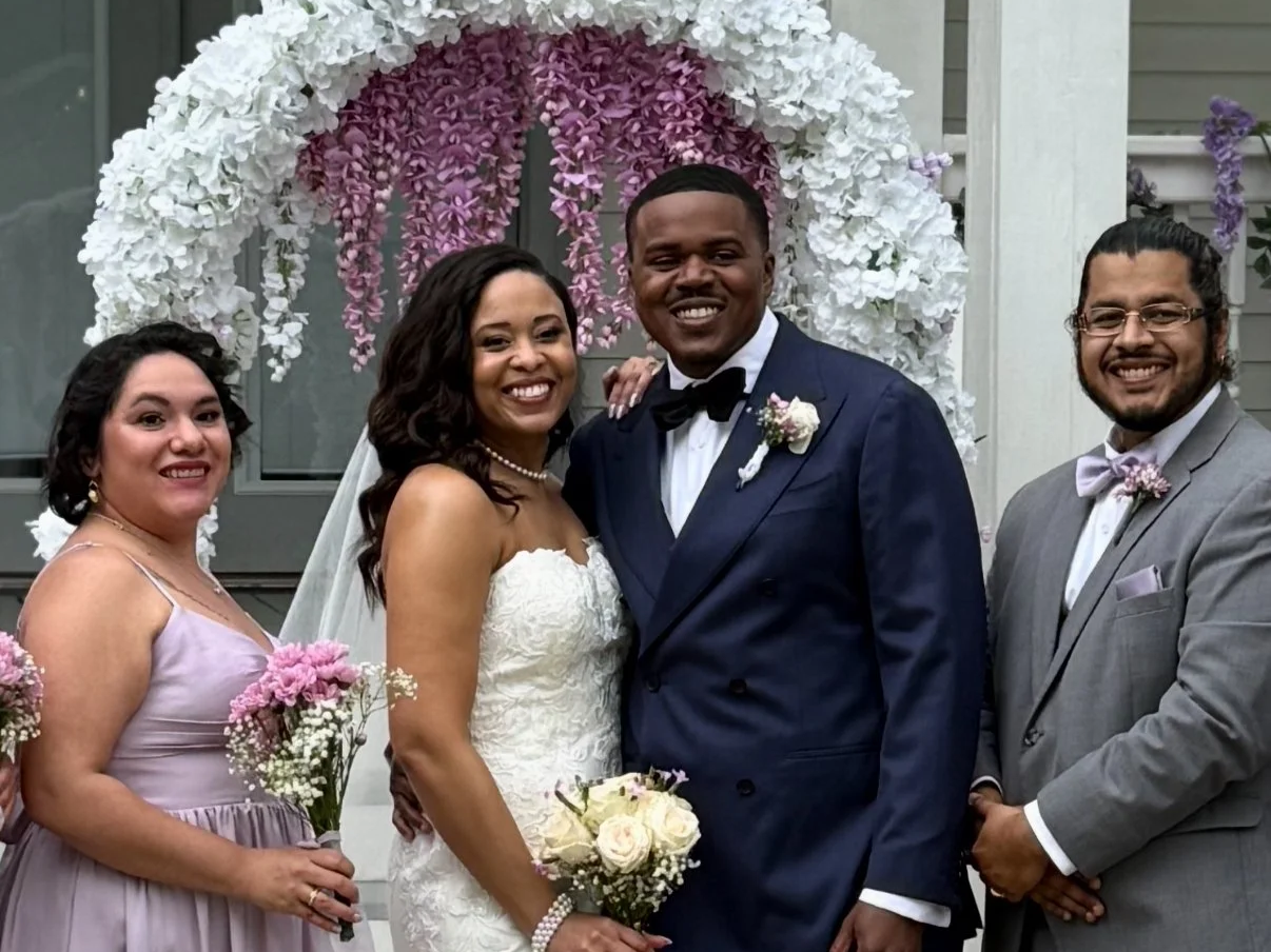 A wedding group portrait with a bride and groom, bridesmaid, and groomsman standing in front of a floral arrangement arch.