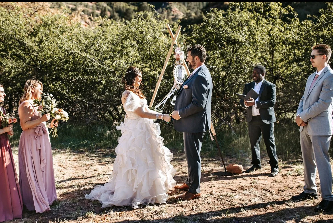 A wedding ceremony outdoors with a bride and groom holding hands in front of a decorated wooden arch, surrounded by bridesmaids and groomsmen, with trees in the background.