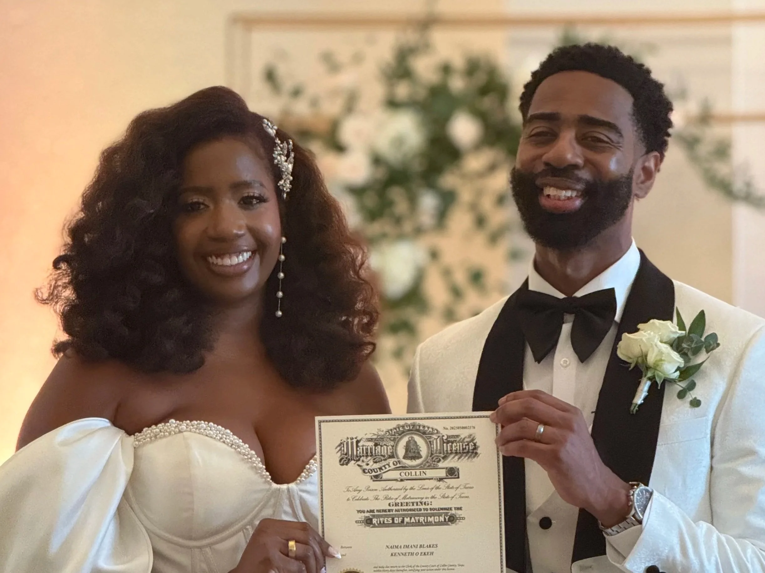 A smiling bride and groom holding a marriage certificate on their wedding day, standing indoors with a blurred floral background.