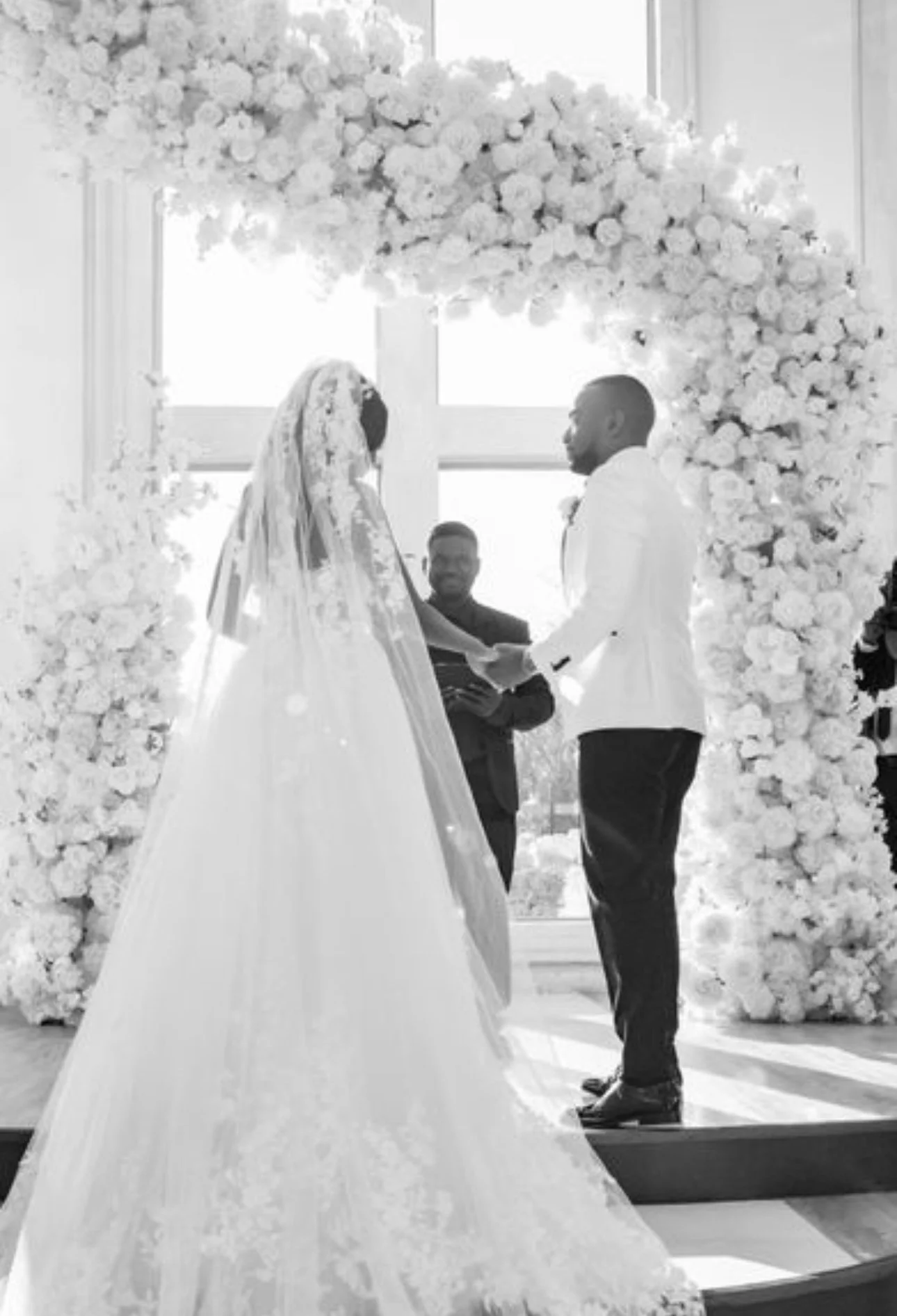 A wedding ceremony with a bride and groom holding hands, standing under a floral arch, with an officiant officiating and a guest observing, inside a bright room with large windows.