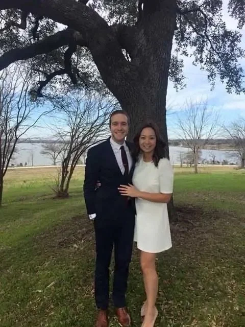 A man and woman standing together outdoors near a large tree, with a lake and blue sky in the background. The man is in a dark suit and the woman is in a white dress.