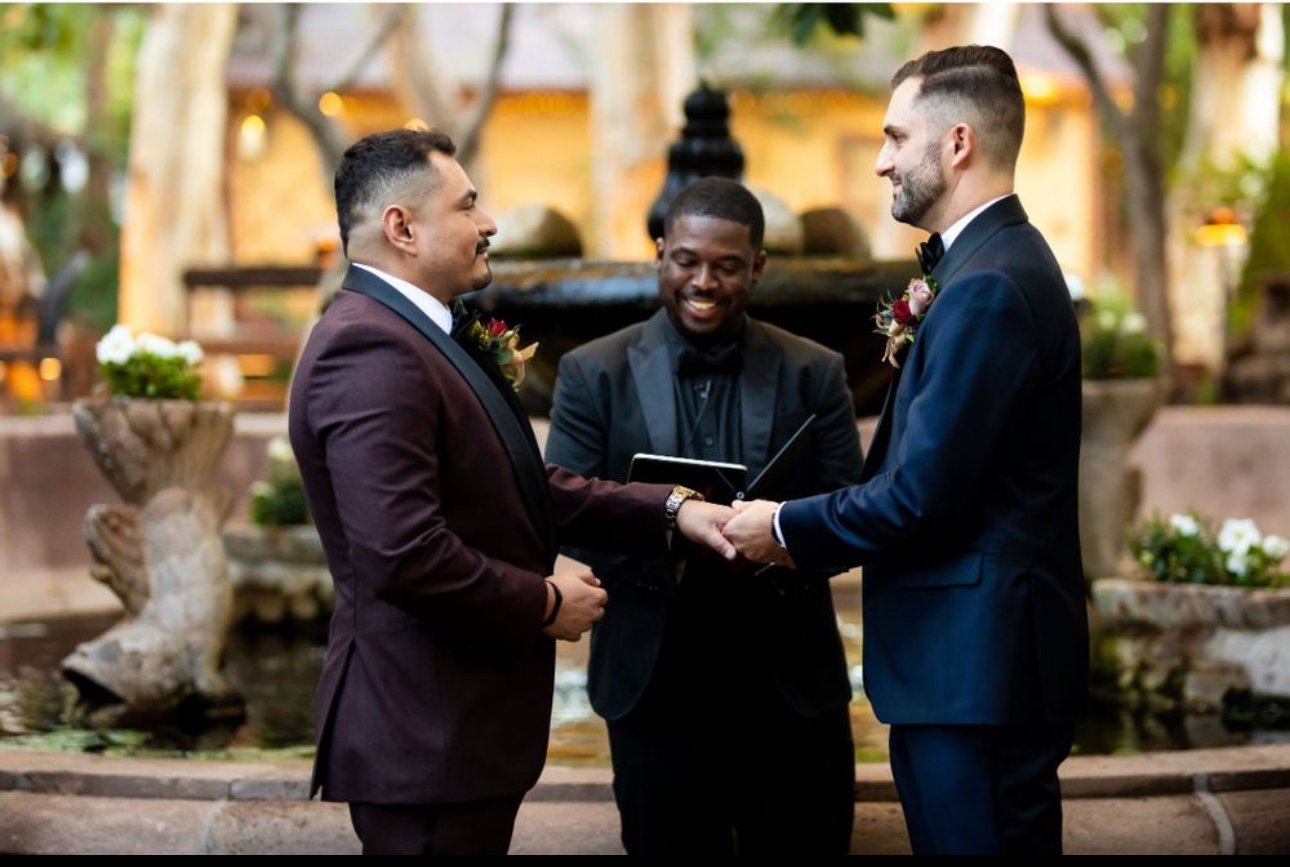 Gay couple exchanging wedding vows outdoors with officiant smiling in the background.