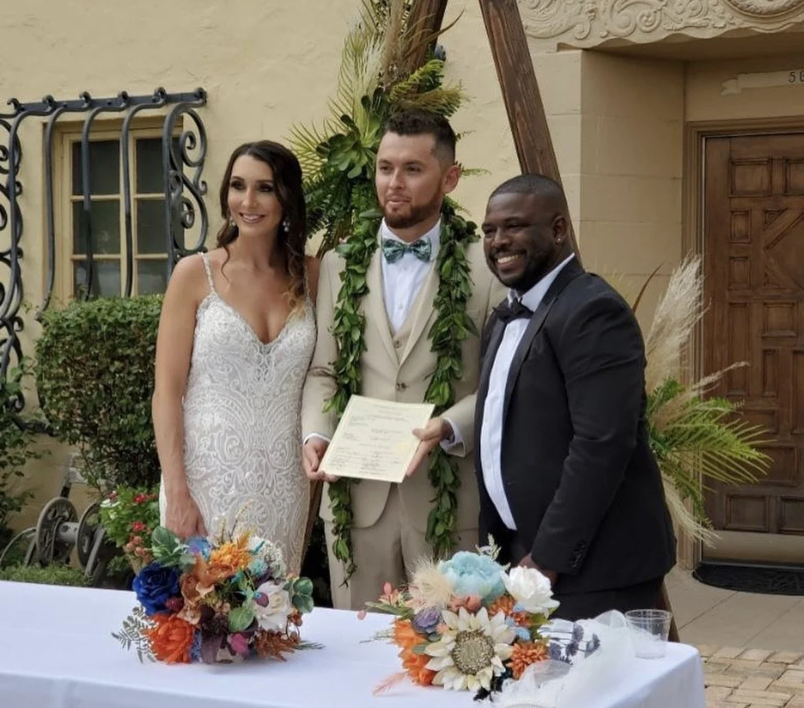 A wedding ceremony with a bride in a white lace gown, a groom in a beige suit with a green leaf lei, and a man in a black tuxedo, holding a document, standing outdoors with decorative plants and a table with colorful flower arrangements.