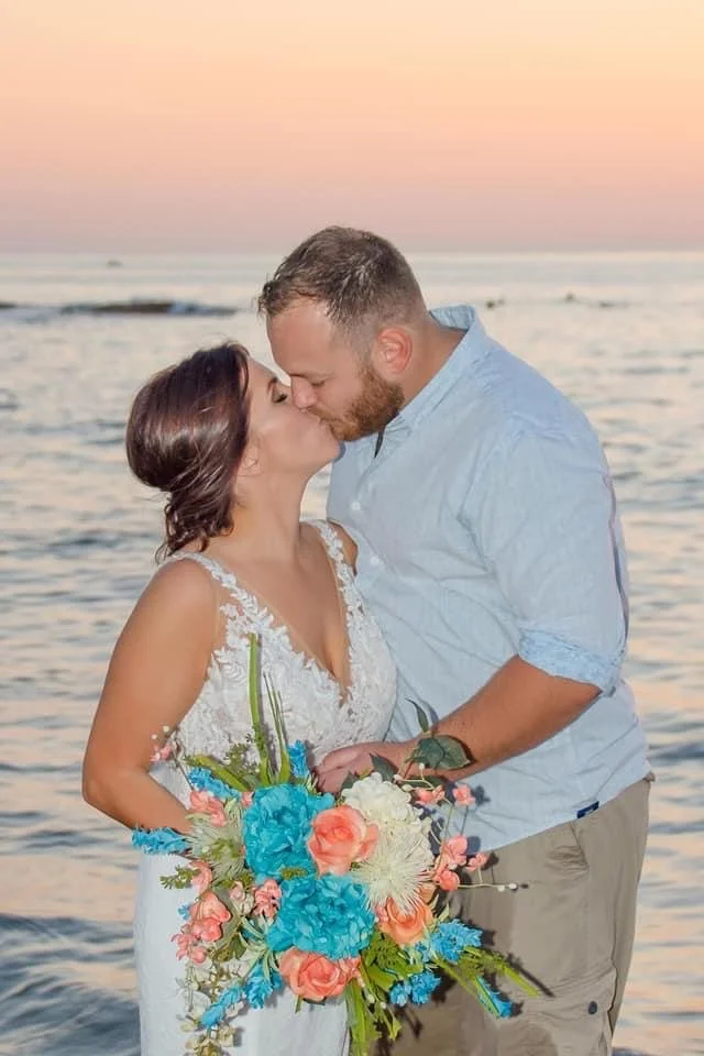 A couple sharing a romantic kiss on the beach during sunset, holding a vibrant bouquet of flowers.
