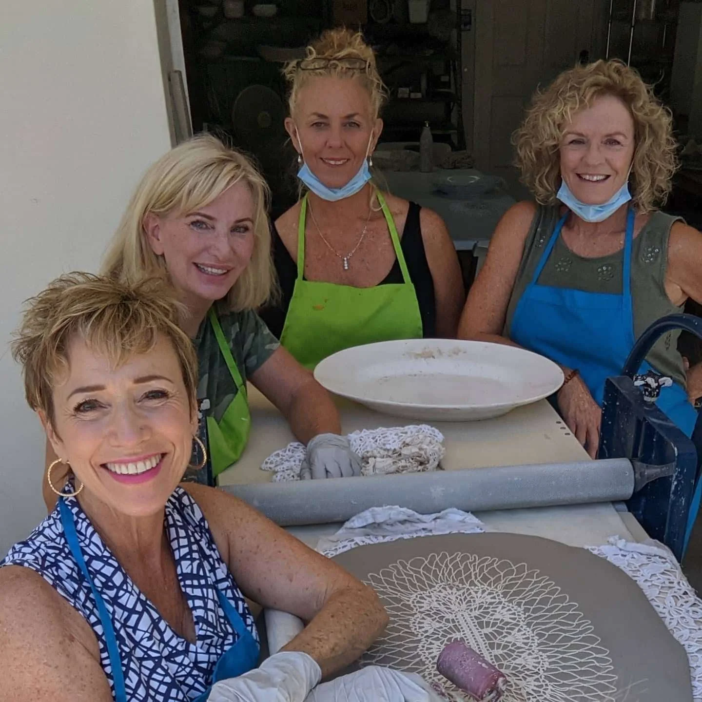 Five women smiling in a kitchen, wearing aprons, with one woman rolling out dough on a table.