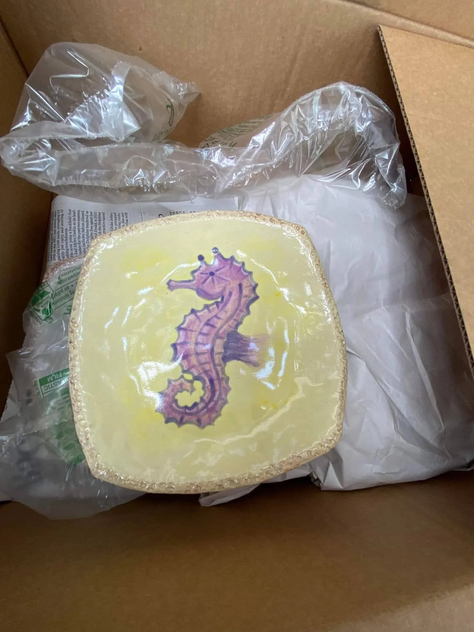 A square piece of cheese with a purple seahorse illustration on top, placed inside a cardboard box with plastic wrapping and newspaper lining.