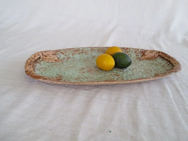 An elongated ceramic or porcelain dish with a textured surface, containing two yellow and one green lemon and placed on a white cloth background.