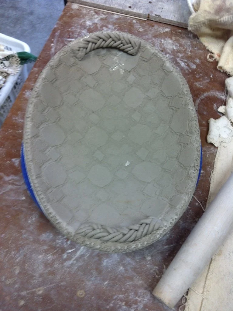Unfinished clay sculpture of an oval-shaped object with decorative braided edges at the top and bottom, resting on a wooden work table.